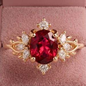 May include: A gold ring featuring a large oval-cut deep red gemstone as the centerpiece, surrounded by smaller marquise and round-cut clear stones arranged in a cluster, set within a gold band and presented inside a dusty pink velvet box.