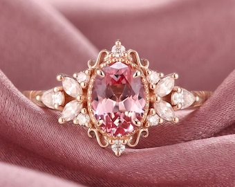 May include: A rose gold ring featuring a central oval-cut pink gemstone surrounded by a decorative filigree setting. The band is accented with marquise-cut and round-cut white stones arranged in a symmetrical cluster on both sides of the center.