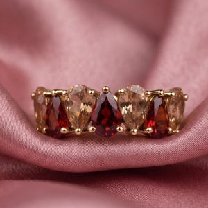 Puede incluir: Un anillo de oro con una serie de piedras preciosas en forma de pera que alternan colores rojo intenso y champán claro. Las piedras están engastadas con garras, creando una fila continua de gemas facetadas alrededor del frente del anillo.