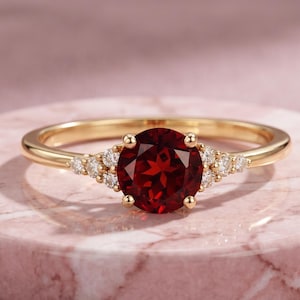 Puede incluir: Un anillo de oro con una piedra preciosa granate redonda de color rojo intenso como piedra central, flanqueada por tres pequeños diamantes redondos transparentes a cada lado, montado en una banda de oro pulido con engaste de garras.