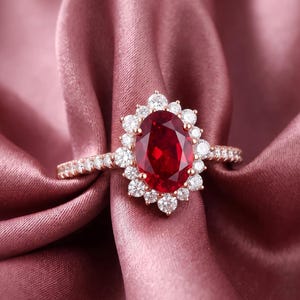 Könnte beinhalten: Ein Ring aus Roségold mit einem großen, oval geschliffenen, tiefroten Edelstein in der Mitte, der von einem Kranz aus kleinen, runden, weißen Steinen umgeben ist. Die Ringschiene ist ebenfalls mit einer Reihe kleiner weißer Steine besetzt.