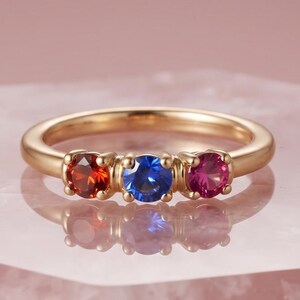 May include: A gold ring featuring three round-cut gemstones set in a row. The stones are arranged with a deep orange-red gem on the left, a vibrant blue gem in the center, and a bright pink-magenta gem on the right, all held by gold prongs.