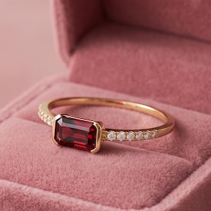 May include: A gold ring featuring a rectangular emerald-cut deep red gemstone set in the center, flanked by a row of small clear round stones along the thin band, presented inside a dusty rose velvet jewelry box with a hinged lid.