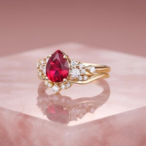 Op de afbeelding: Een gouden ring met een centrale peervormige rode edelsteen omringd door een cluster van kleine ronde witte diamanten. De band is gemaakt van gepolijst geelgoud en de zetting bevat meerdere accentstenen in een bloemvormig patroon.