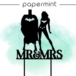 Op de afbeelding: Zwarte silhouet van Batman en Wonder Woman figuren met de tekst "MR&MRS" op een witte achtergrond. De figuren staan op een taarttopper met drie stokjes.