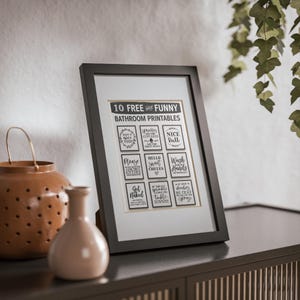 Puede incluir: Una impresión enmarcada con el texto "10 FREE and FUNNY BATHROOM PRINTABLES" y varias frases humorísticas. El marco negro se encuentra sobre una superficie oscura, con objetos decorativos en primer plano.