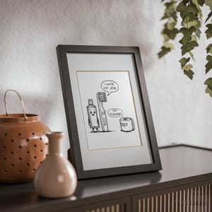 Puede incluir: Una impresión de dibujos animados en blanco y negro enmarcada que representa un cepillo de dientes, pasta de dientes y papel higiénico con bocadillos. Los bocadillos dicen "I hate my job" y "Oh please!"