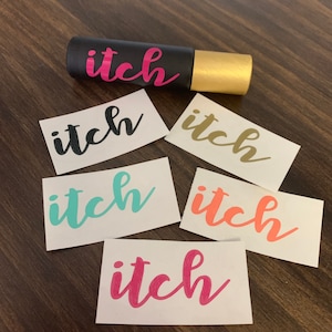 May include: Six white stickers with the word "itch" written in different colors: black, gold, teal, orange, and pink. A black roller bottle with a gold cap also has the word "itch" written on it.