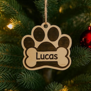 May include: Wooden paw print Christmas ornament with the name "Lucas" on a bone-shaped accent. The ornament is hanging from a twine string against a backdrop of a Christmas tree with a red ornament.