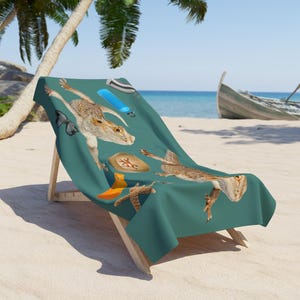 May include: Teal beach towel featuring a whimsical design of a lizard with beach-themed accessories. The towel includes a lizard illustration, sunglasses, a hat, and a blue floating device. The towel is draped over a wooden beach chair on a sandy beach.