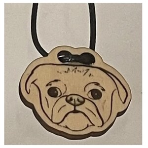 May include: A wooden pendant necklace featuring a pug dog design. The pendant is light brown with dark brown etched details and a black cord. The pug's face is detailed with eyes, nose, and wrinkles. The pendant has a small black bow.