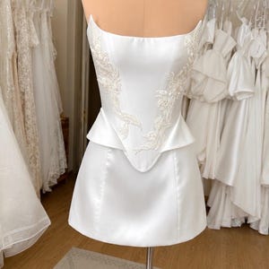 May include: A white strapless corset-style top with a peplum waist and a short skirt. The bodice features intricate white floral embroidery and beadwork detailing along the front panels, crafted from a smooth, satin-like fabric with a structured fit.