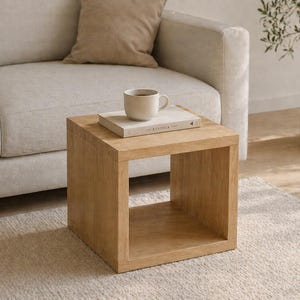 Puede incluir: Una mesa auxiliar cuadrada de madera clara. Un libro y una taza beige están encima. La mesa está frente a un sofá de color claro y una planta en maceta. Dimensiones aproximadas 40 cm x 40 cm x 40 cm.
