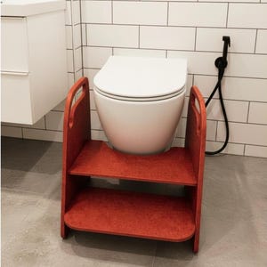 May include: A two-step stool made of reddish-brown material with a textured finish. The stool features two horizontal platforms and two side panels with integrated handle cutouts, positioned directly in front of a white wall-mounted toilet.