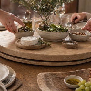 Könnte beinhalten: Ein Holztisch mit einem hölzernen Lazy Susan, beladen mit Schalen mit Oliven, Nüssen und Grünzeug. Ein kleiner Käselaib liegt auf einem Holztablett. Weingläser und Teller sind um den Tisch herum aufgestellt, was auf ein Essen hindeutet.