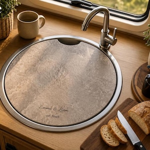 Könnte beinhalten: Eine runde Spülenabdeckung in Steinoptik mit Griffmulde und der Gravur "Emma & Liam on tour". Die Abdeckung liegt bündig in einem runden Edelstahl-Spülbecken unter einem silbernen Metallwasserhahn auf einer Arbeitsplatte aus Holz.