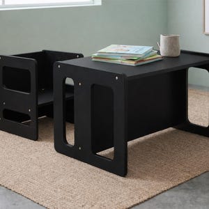 Op de afbeelding: Een zwarte kinderstoel en -tafelset. De tafel heeft een rechthoekig blad met uitgesneden zijkanten en een massieve voorkant. De stoel heeft een vierkante rugleuning en zitting met uitgesneden zijkanten. Een stapel boeken en een kleine mok staan op de tafel.