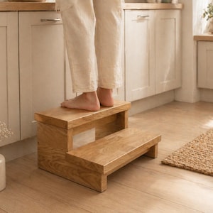 Puede incluir: Un taburete de madera de dos peldaños con un acabado de veta de madera marrón claro natural. El taburete cuenta con un peldaño superior rectangular y un peldaño inferior, proporcionando una plataforma estable para alcanzar lugares altos.