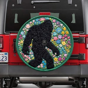 Op de afbeelding: Een rode Jeep met een zwarte reservewielhoes met een zwart Bigfoot-silhouet. De hoes is omgeven door kleurrijke bloemen op een groene achtergrond. De kentekenplaat luidt "SAM123".