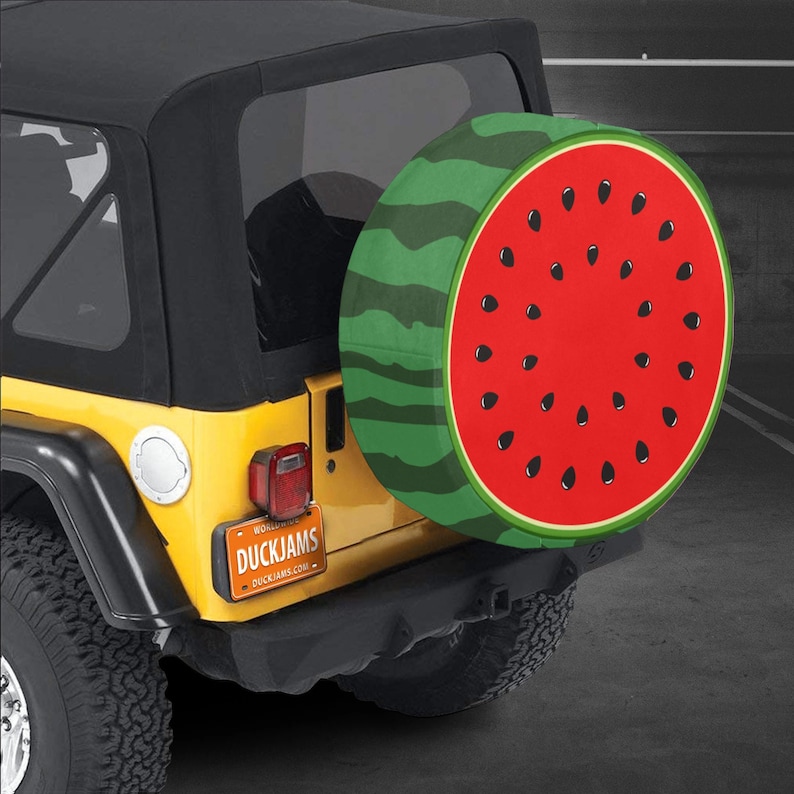 May include: A circular spare tire cover featuring a vibrant red watermelon flesh pattern with black seeds on the face and a green rind pattern on the side, mounted on the rear of a yellow vehicle with a license plate reading DUCKJAMS.