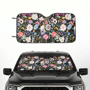 May include: A rectangular car windshield sun shade featuring a dense botanical print of various flowers including pink peonies, white dahlias, and blue hydrangeas against a black background, shown both flat and installed in a black vehicle.