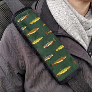 May include: A dark green seat belt cover featuring a repeating pattern of multi-coloured trout fish. The rectangular fabric accessory has black trim along the edges and is secured over a grey seat belt strap, providing a cushioned surface.