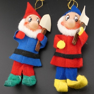 May include: Two felt Christmas ornaments shaped like gnomes, each holding a small wooden shovel. One gnome is wearing a red hat, blue pants, and a red and green outfit. The other gnome is wearing a blue hat, red pants, and a blue and yellow outfit.