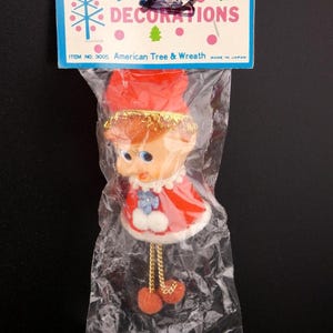 May include: Vintage Christmas ornament of a doll in a red dress with white trim, a red hat, and gold hair. The ornament is in a clear plastic bag with a blue and pink tag that reads "HOLIDAY DECORATIONS".