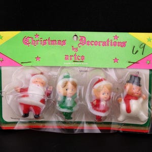 May include: Vintage Christmas decorations by Artco, featuring four plastic ornaments. The set includes Santa Claus, a girl in a green dress, a girl in a red dress, and a snowman. The packaging is green and pink with the words "Christmas Decorations".