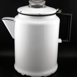 May include: White enamel coffee pot with a black handle and a glass lid. The pot has a black trim around the top and bottom.