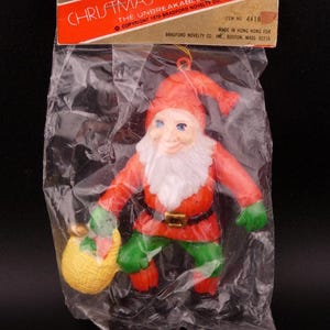 May include: Vintage Christmas decoration of a gnome in a plastic bag. The gnome wears a red hat and jacket, green pants, and a white beard. It holds a yellow sack. The packaging reads "CHRISTMAS DECORATIONS" and "BRADFORD'S".