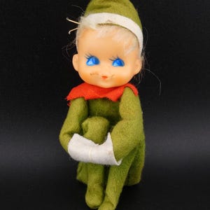 May include: A vintage elf doll with a green felt outfit, a red collar, and a white headband. The doll has a painted face with blue eyes and blonde hair. The elf is sitting with its arms crossed.