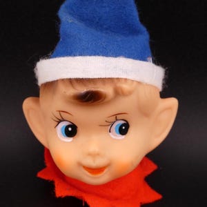 May include: A vintage elf head with a blue felt hat trimmed with white. The elf has a painted face with blue eyes, rosy cheeks, and a red felt collar. The background is black.