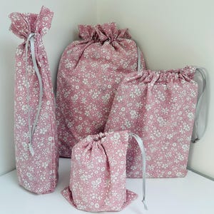 May include: A set of pink floral fabric drawstring bags in various sizes. Each bag features a white floral pattern and a grey ribbon drawstring. The bags are displayed against a white backdrop.