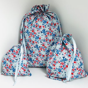 May include: Three drawstring bags in different sizes, adorned with a floral pattern of red, blue, and pink flowers on a pale blue fabric. Each bag has a light blue ribbon drawstring. The bags are displayed in a corner.