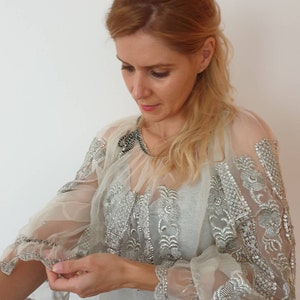 Op de afbeelding: Een doorschijnend wit blouse met zilveren borduurwerk en pailletten. De blouse heeft lange mouwen met een gerimpelde manchet en een ronde hals.
