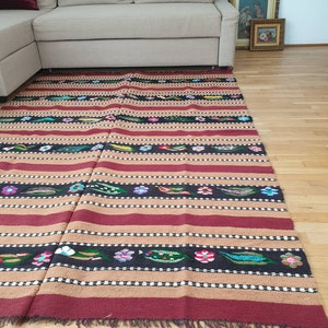 May include: A rectangular, hand-woven rug with a striped pattern in shades of red, tan, and black. The rug features floral and leaf motifs in various colours, including pink, blue, and green. The rug has a fringed edge and is placed on a hardwood floor.