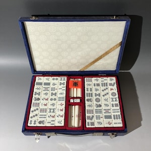 May include: A mahjong set in a blue fabric-covered carrying case with a white patterned interior lining. The set includes white tiles with black, red, and blue symbols, along with white counting sticks and dice arranged in red velvet-lined compartments.