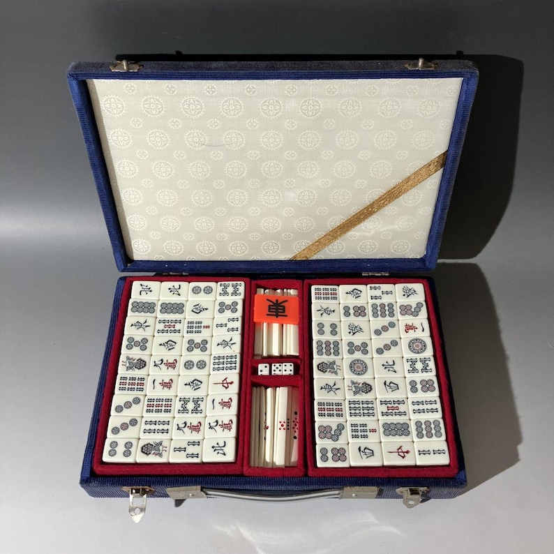 May include: A mahjong set in a blue fabric-covered carrying case with a white patterned interior lining. The set includes white tiles with black, red, and blue symbols, along with white counting sticks and dice arranged in red velvet-lined compartments.