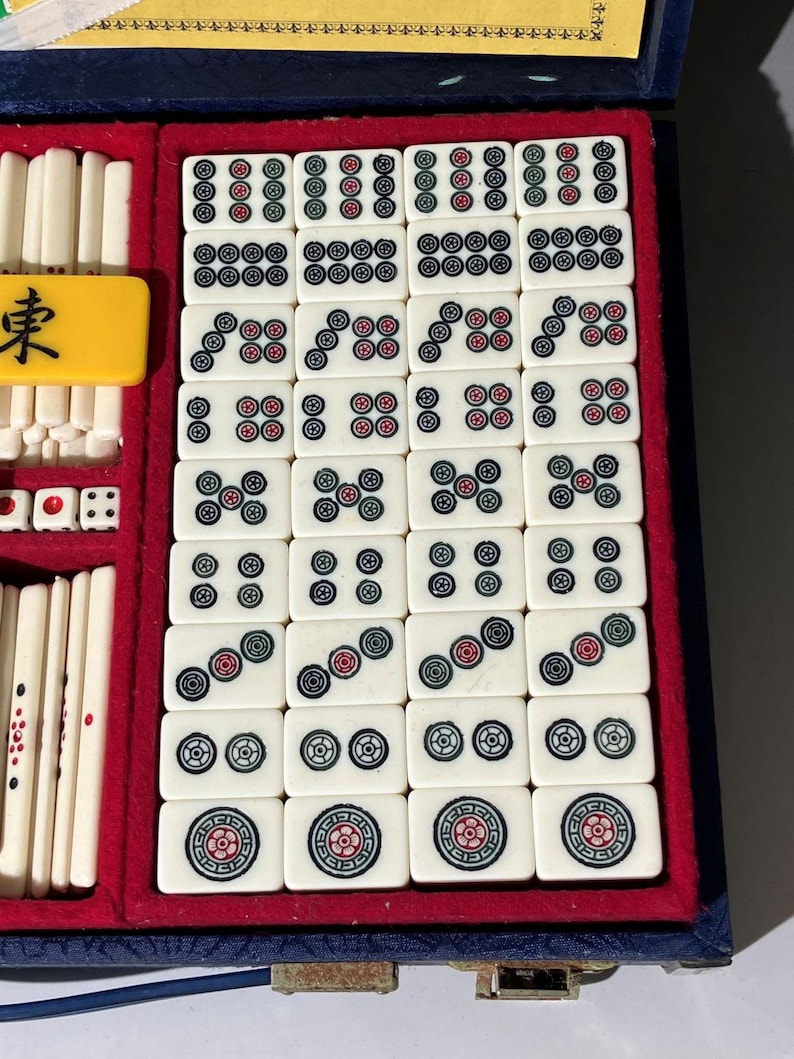 May include: A vintage Mahjong set in a blue velvet case with red lining. The ivory-colored tiles feature black, red, and green symbols and numbers. Includes dice and a yellow tile with a black character.