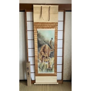 Pode incluir: Uma pintura japonesa tradicional em rolo que retrata um tigre em uma paisagem montanhosa. O tigre é pintado em tons de marrom e laranja, com uma barriga branca. O fundo é um azul e verde suave, com toques de ouro. O rolo é montado em um rolo de madeira e tem uma borda decorativa.