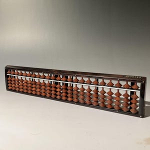 Puede incluir: Un ábaco de madera vintage con filas de cuentas marrones. El ábaco tiene un marco marrón oscuro y varillas de metal blanco. Las cuentas están dispuestas en columnas, sobre un fondo neutro.