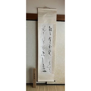 以下が含まれることがあります： クリーム色の紙に黒い書道が施された、縦長の日本の掛け軸。掛け軸は吊り下げられており、下部にはテキストが書かれた木箱があります。背景は明るい色の壁です。