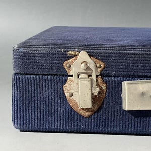 May include: A rectangular storage box covered in dark blue corduroy fabric. The box features a silver-toned metal latch with visible signs of oxidation and wear, secured to the front panel. A metallic handle is partially visible on the right side.