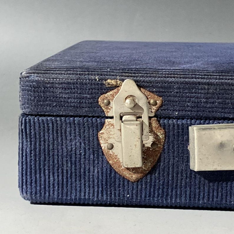 May include: A rectangular storage box covered in dark blue corduroy fabric. The box features a silver-toned metal latch with visible signs of oxidation and wear, secured to the front panel. A metallic handle is partially visible on the right side.
