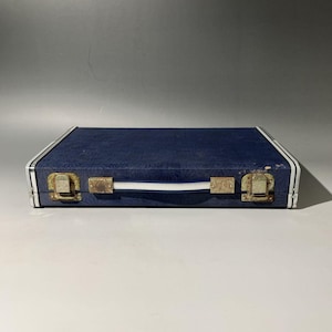 May include: A rectangular, navy blue case with a textured surface. The case has a white handle and white stripes along the edges. Two tarnished metal clasps secure the case.