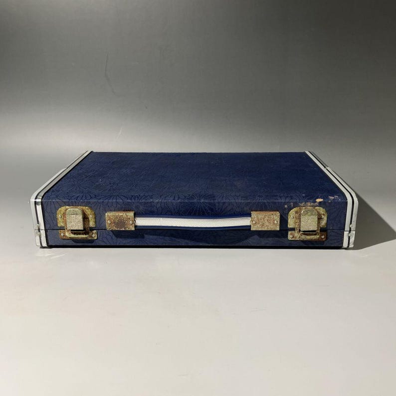 May include: A rectangular, navy blue case with a textured surface. The case has a white handle and white stripes along the edges. Two tarnished metal clasps secure the case.