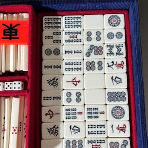 May include: A Mahjong set featuring off-white tiles with black, red, and blue engraved symbols, numbers, and characters. The tiles are organized in a blue velvet-lined case with red dividers, including a bright orange marker with the black character 東.