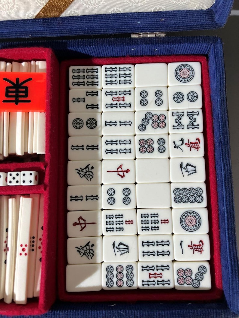 May include: A Mahjong set featuring off-white tiles with black, red, and blue engraved symbols, numbers, and characters. The tiles are organized in a blue velvet-lined case with red dividers, including a bright orange marker with the black character 東.