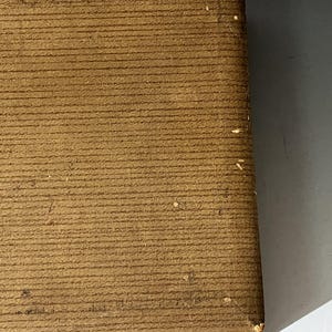 Puede incluir: Una caja rectangular de color marr&oacute;n con una superficie texturizada y acanalada. La caja presenta una esquina visible con un ligero desgaste y peque&ntilde;as rozaduras claras. Un mecanismo de cierre met&aacute;lico es parcialmente visible en el borde.